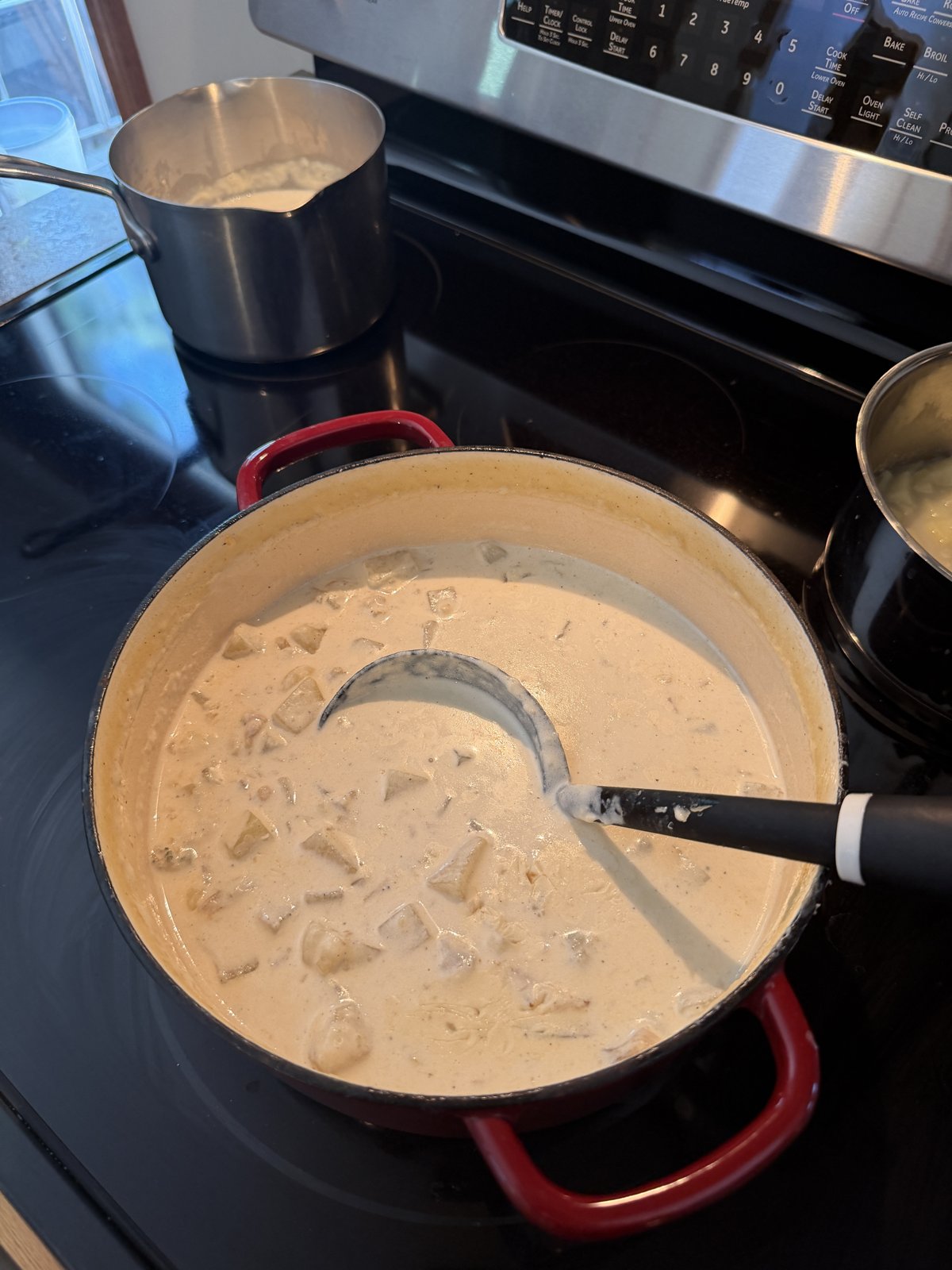 A red Dutch oven on the stove, full of creamy clam chowder with bite-sized potato pieces, a black ladle resting in the pot