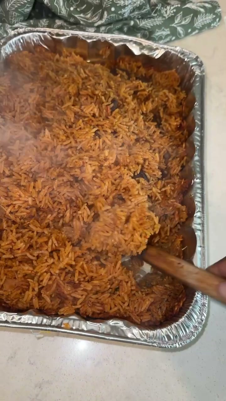 A foil pan of steaming orange-red jollof rice with a wooden spoon digging in